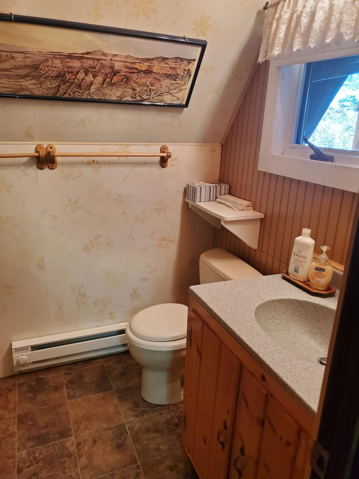 A compact bathroom features natural wood accents and a light-colored countertop. The space includes a toilet, a sink with toiletries, and a shelf displaying neatly stacked items. A window allows natural light, and floral-patterned wallpaper adds a soft touch to the walls.