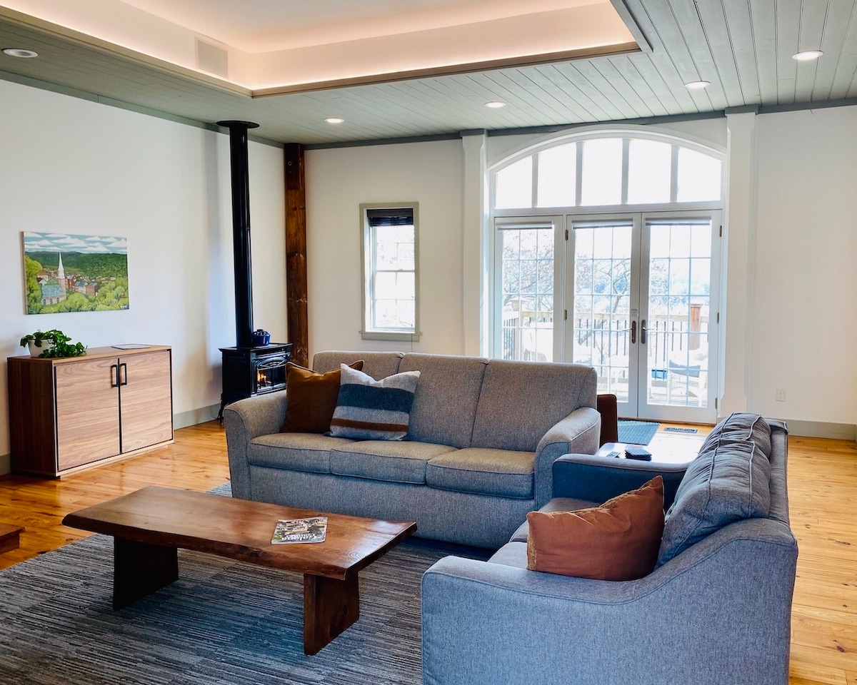 The spacious living area features hardwood floors and large windows that allow natural light to fill the room. A comfortable gray sofa and loveseat are complemented by a wooden coffee table. A gas fireplace provides warmth, while French doors open to a private deck with scenic views.