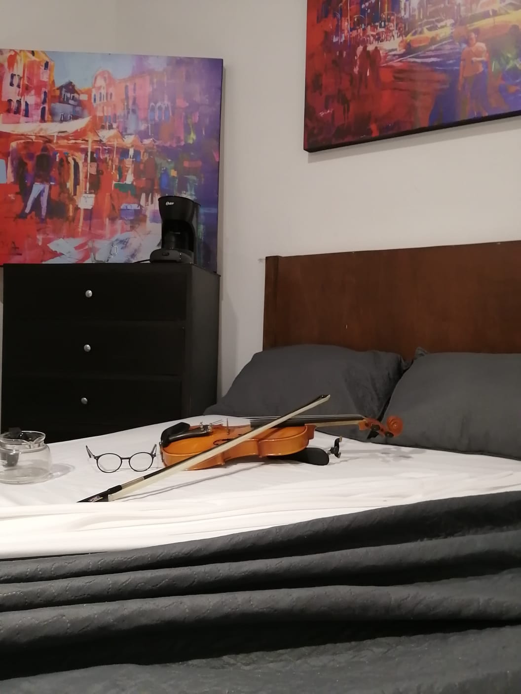 A cozy sleeping area features a spacious bed with gray linens and an assortment of items on top, including a violin, a pair of eyeglasses, and a small ashtray. A coffee maker and a tall dresser are visible in the background, complemented by colorful wall art.