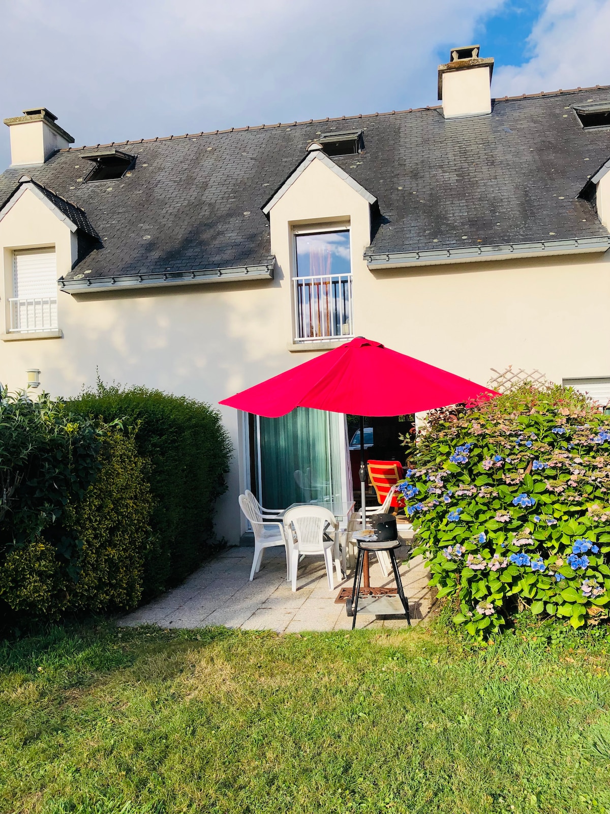 A charming outdoor space is presented, featuring a red umbrella providing shade over a small table and four chairs. Lush greenery, including hydrangeas, frames the patio area, while the exterior of the house showcases two upper windows and a glass door leading inside.