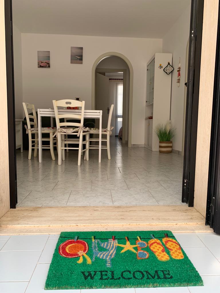 The image features an inviting entryway with a colorful welcome mat. A spacious dining area can be seen through the doorway, furnished with a large wooden table surrounded by light-colored chairs. Natural light enters through a nearby window, illuminating the room's neutral tones.