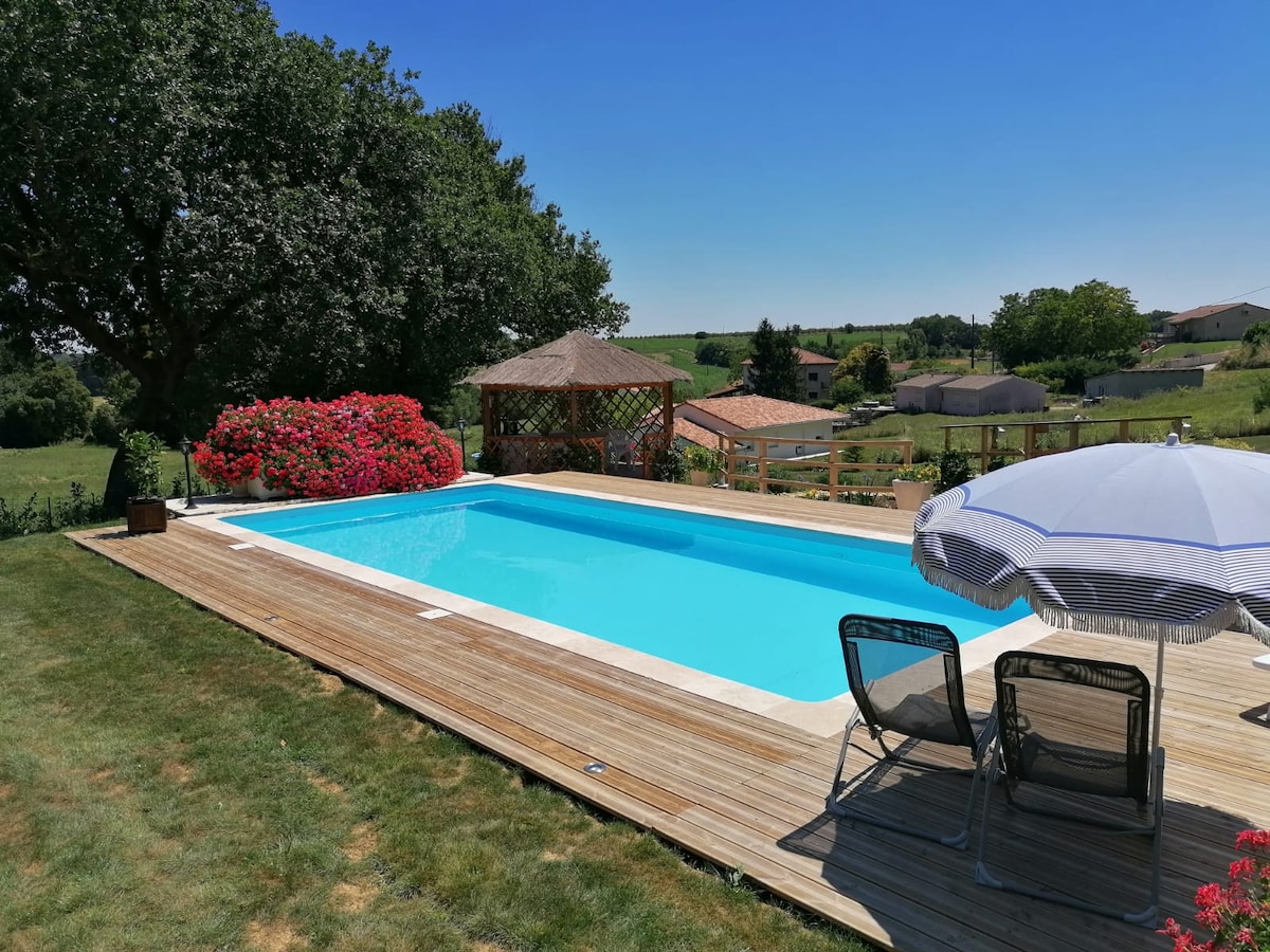 A private swimming pool is surrounded by a wooden deck, featuring lounge chairs and an umbrella for shade. Lush greenery and vibrant flowers frame the pool area, while a gazebo is visible in the background, offering additional outdoor relaxation space.