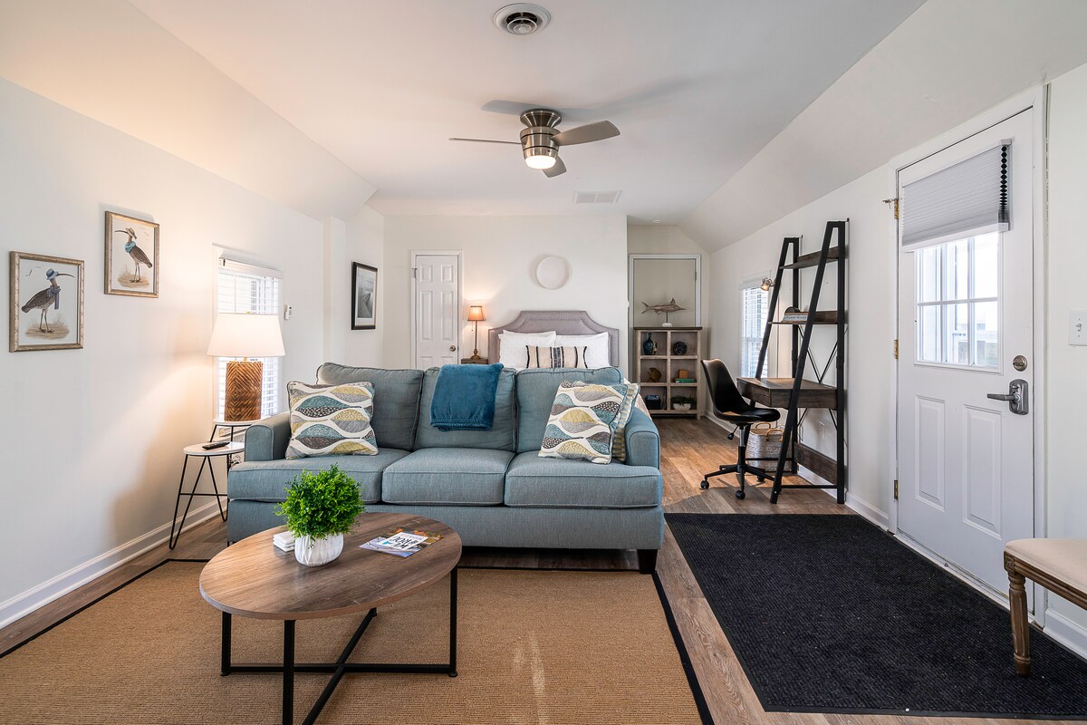 A bright and airy efficiency apartment is showcased, featuring a cozy seating area with a blue sofa and a round coffee table. A desk is placed against the wall, and a door provides private entrance access. Natural light floods the space through the windows, highlighting the decorative wall art.