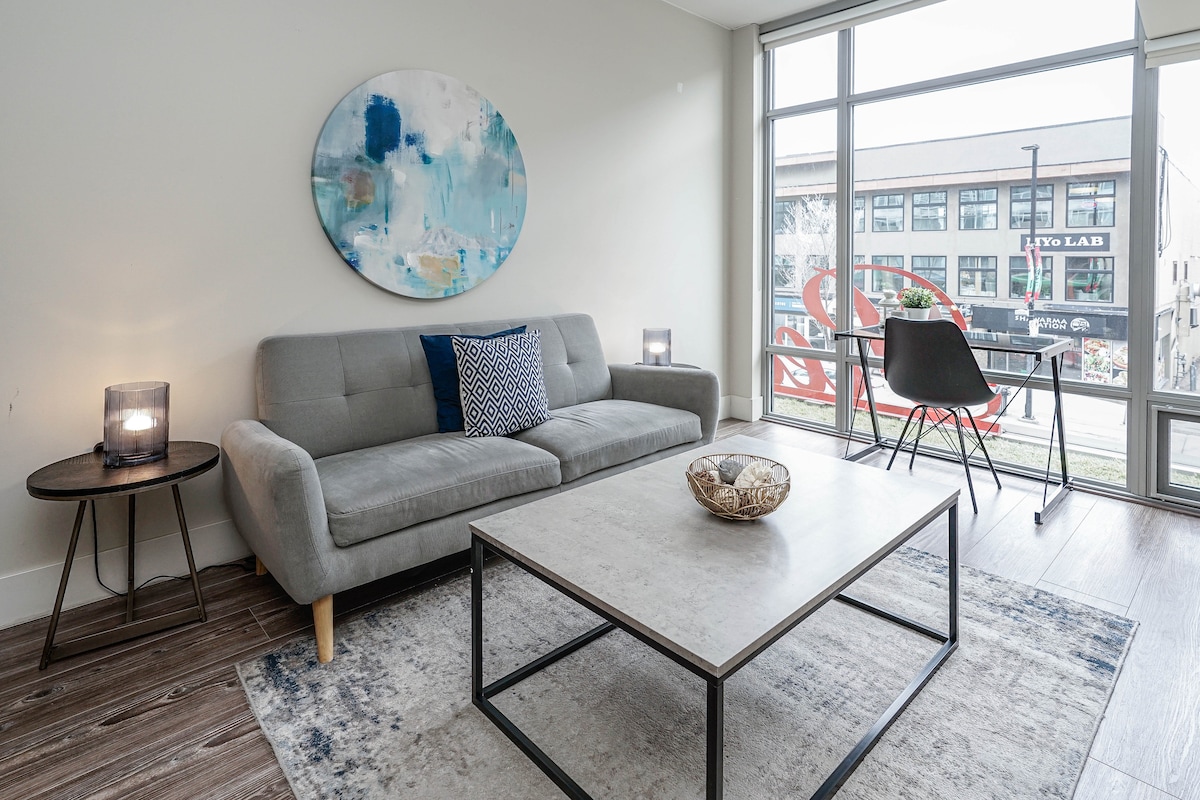 A bright living room is showcased with large windows allowing natural light. The space features a modern gray sofa with accent pillows, a low coffee table, and a decorative bowl. Soft lighting from two side tables complements the contemporary decor.