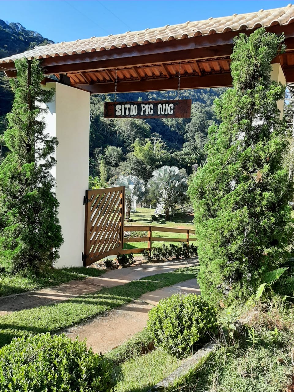A welcoming entrance features a wooden gate set within a stone pathway. Lush green shrubs flank either side, leading to a garden area with vibrant vegetation and tall trees, creating a serene and inviting atmosphere.