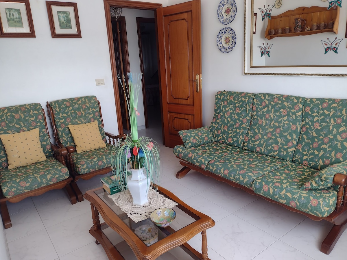 A living area is furnished with two patterned armchairs and a matching sofa. A wooden coffee table occupies the center, adorned with a decorative cloth and a vase of greenery. Walls are decorated with framed art and ceramic plates, adding character to the space.