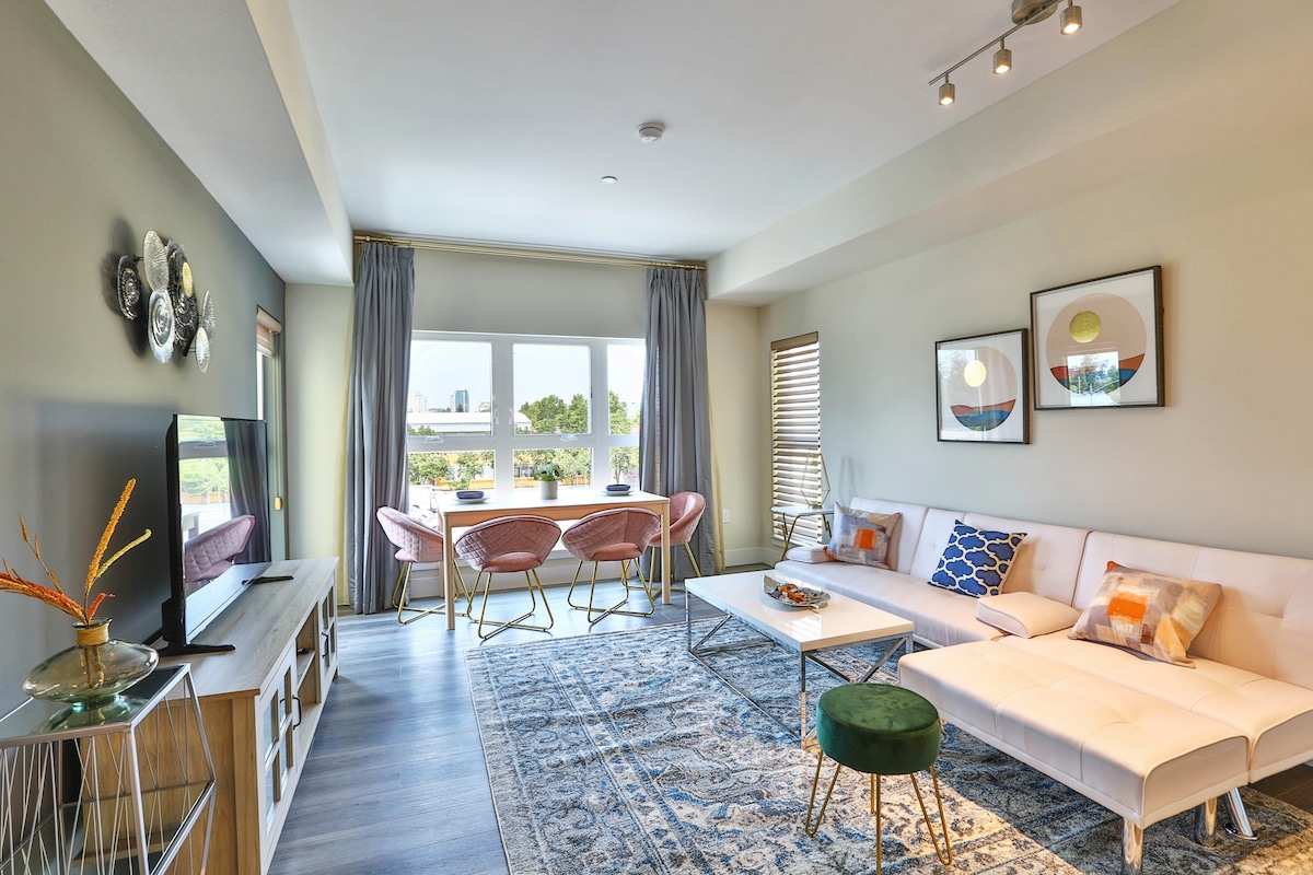 A bright living area features a white sectional sofa and a coffee table atop a decorative area rug. Two framed artworks adorn the wall, while large windows invite natural light, showcasing outdoor views. Accent chairs in soft pink add a stylish touch to the space.