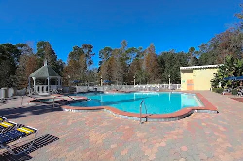 A community pool area is surrounded by tall trees, providing a sense of calm. A gazebo and lounge chairs are visible near the pool, which features a curved shape and stair access. The sunny sky adds to the inviting atmosphere.
