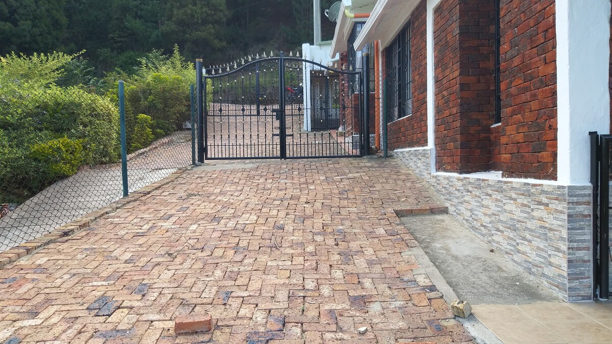 A gated entrance opens onto a paved driveway, bordered by greenery. The structure features a combination of brick and stone materials, with windows adorned by black grilles. The surrounding area is well-maintained, creating a welcoming approach to the property.