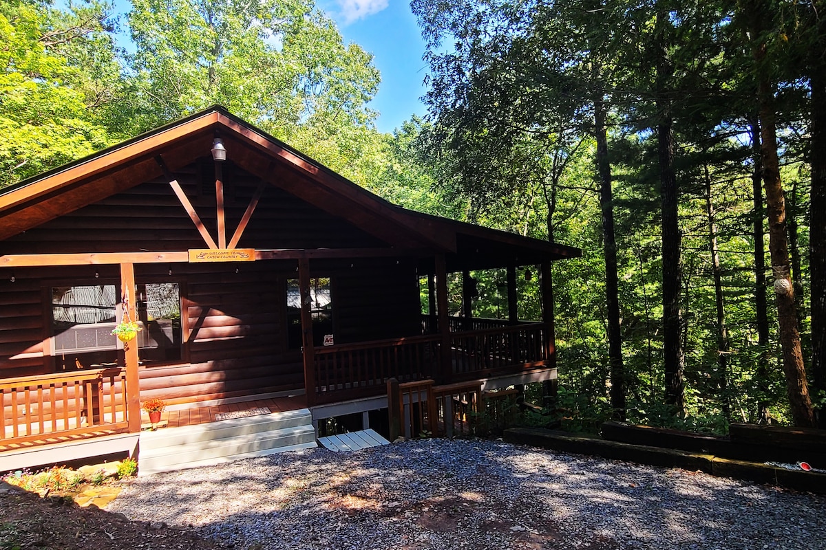 The exterior of a cabin is shown, surrounded by lush green trees. A spacious porch with wooden railings is visible, providing an inviting entrance. Gravel is laid in the parking area, accommodating vehicles with easy access to the steps leading to the porch.