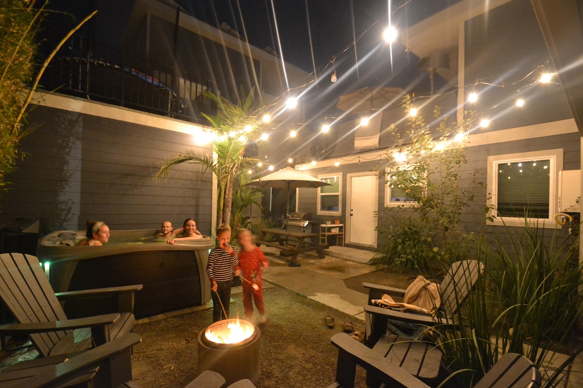 A cozy courtyard is illuminated by string lights on a clear night. A six-person hot tub is positioned next to a seating area featuring wooden adirondack chairs. In the background, a picnic table can be seen, surrounded by greenery and soft outdoor lighting.