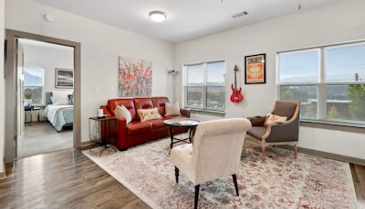 A comfortable living area is presented, featuring a red sofa and a beige armchair beside a round table. Art pieces adorn the walls, while large windows allow ample natural light. A doorway leads to another room, providing a sense of openness.