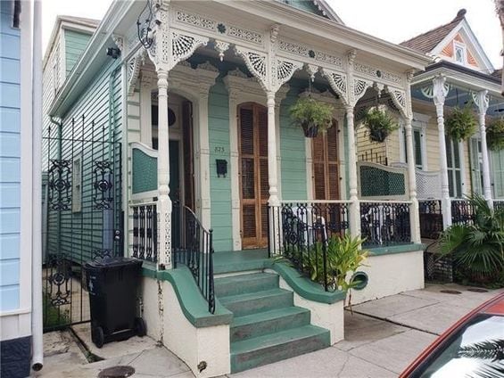 A charming exterior view of a historic home features intricate wooden detailing on the porch and balustrades. Green steps lead up to a door framed by wooden shutters. Potted plants add greenery to the welcoming entrance, complemented by a light blue facade.