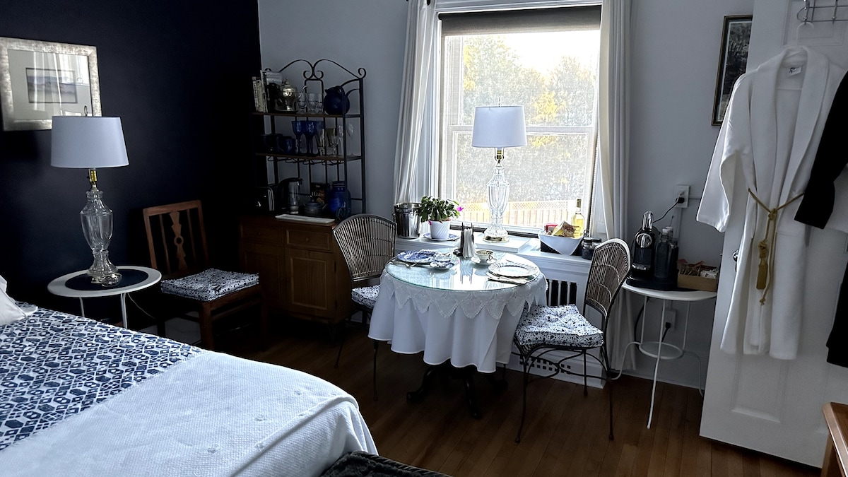 The cozy room features a spacious bed and a round dining table surrounded by four chairs. Natural light enters through a window, illuminating the space. A small kitchenette area is visible, along with a closet displaying housecoats.
