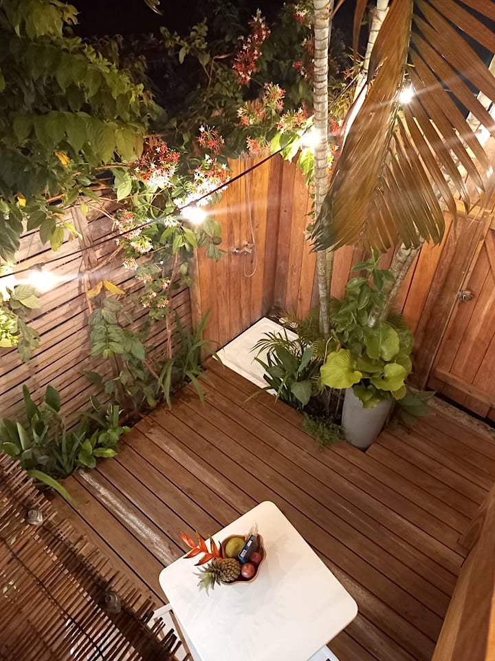 An outdoor shower area features wooden decking surrounded by lush greenery and potted plants. A small table is adorned with fresh tropical fruits, while lights are strung above, creating a cozy ambiance. Nature-inspired elements convey a sense of tranquility in the space.