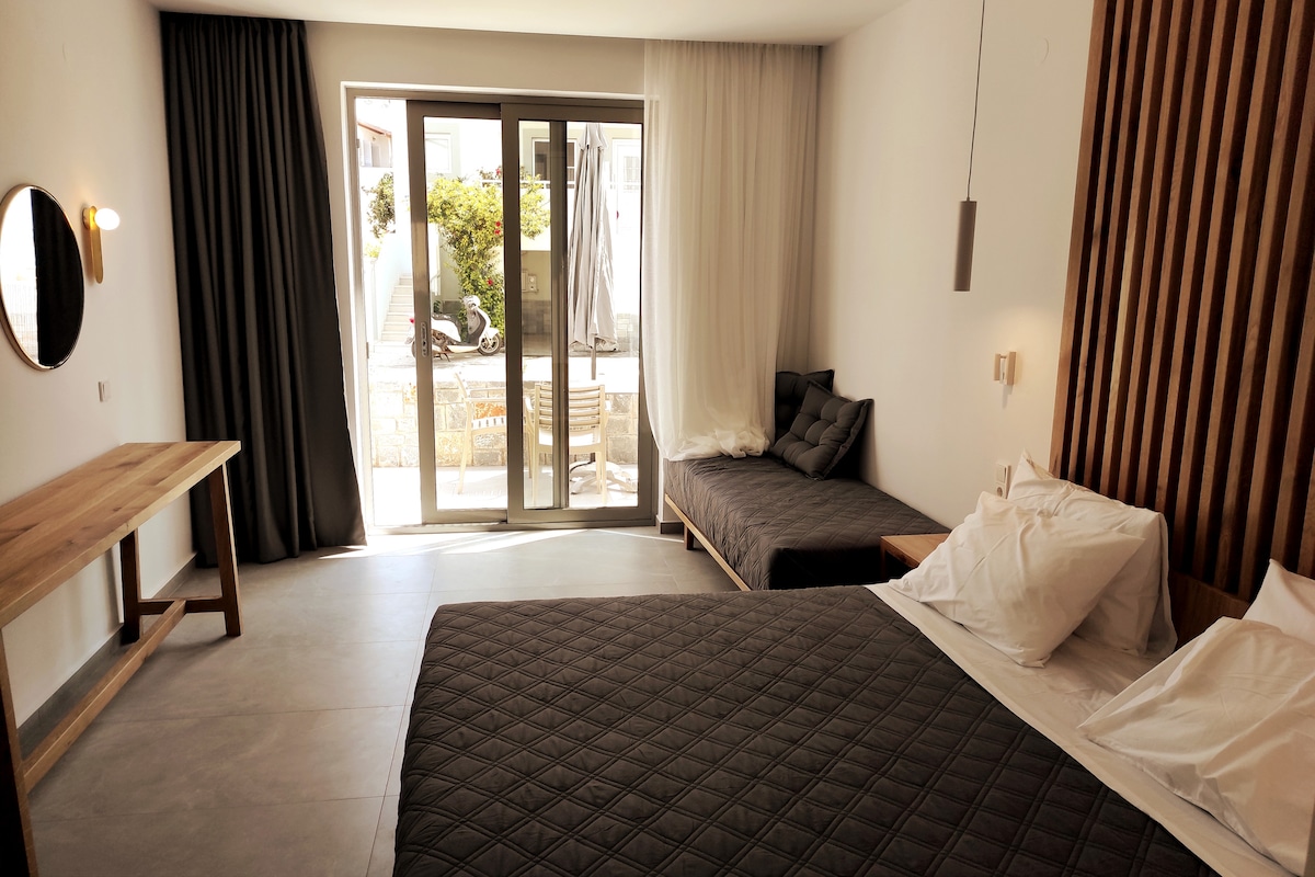 A spacious room features a large bed covered with a dark quilt and light pillows. A second, smaller bed is placed against the wall. Sliding glass doors lead to an outdoor seating area, with natural light illuminating the modern decor.