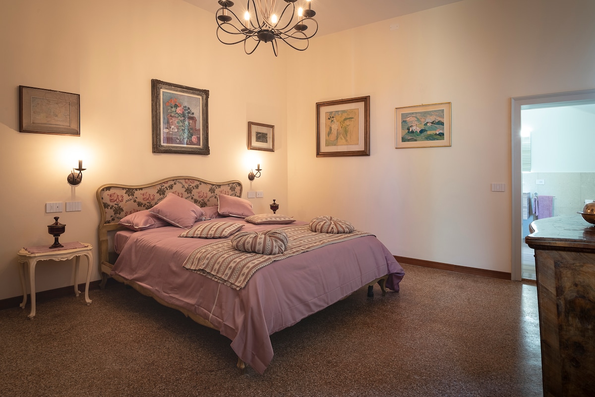 A spacious bedroom is featured, showcasing a decorative bed with soft lavender bedding and several accent pillows. Framed artwork adorns the walls, while wall sconces provide gentle lighting. A doorway leads to an adjoining bathroom, contributing to the room's overall elegance.