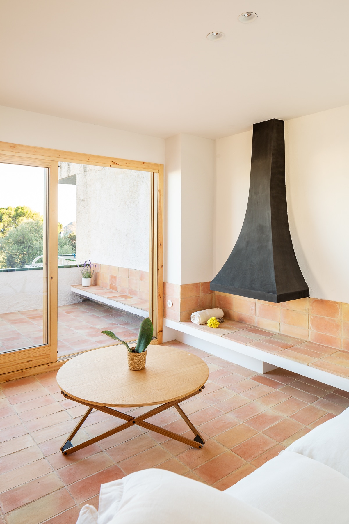 A bright living area is featured, showcasing a wooden coffee table placed on terracotta tiles. A minimalist black chimney is mounted on the wall, enhancing the space. Large glass doors provide access to an outdoor area, inviting natural light into the room.