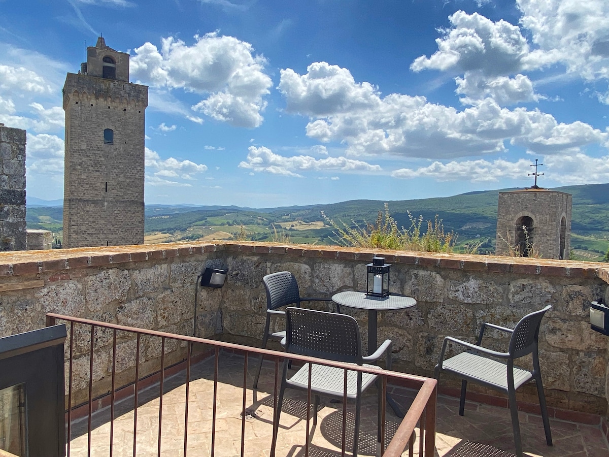 A scenic terrace showcases views of rolling hills and historic towers under a vibrant sky. Two chairs and a small table are positioned for outdoor relaxation, while stone walls provide a rustic backdrop to the peaceful setting.