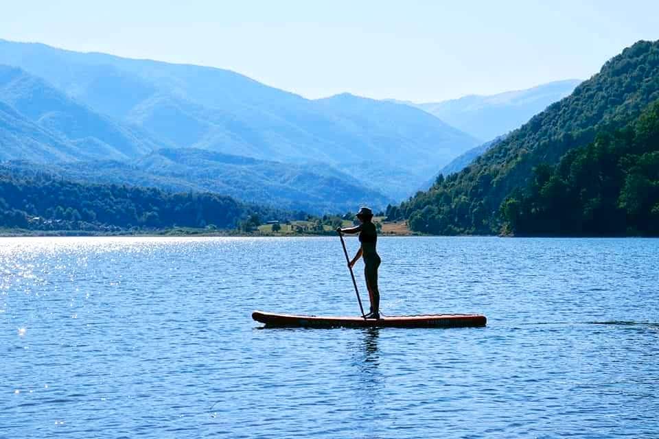 A paddleboarder is seen gliding across the calm water, surrounded by rolling mountains under a clear blue sky. The landscape features soft hills in the background, creating a serene atmosphere. Sunlight reflects off the water's surface, enhancing the tranquil scene.