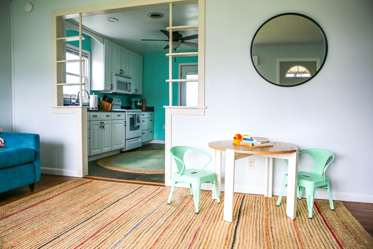 A cozy corner is presented with a circular rug and a light-colored table surrounded by two green chairs. A round mirror hangs on the wall, reflecting the airy kitchen space visible in the background, featuring smooth cabinetry and bright accents.