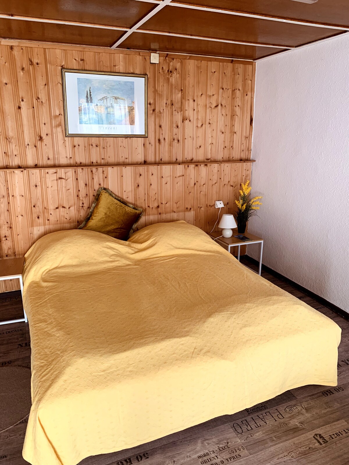 A sleeping area features a spacious bed covered with a yellow bedspread. The wooden paneling adds warmth to the room, complemented by a small bedside table with a lamp and a decorative vase. A framed artwork hangs on the wall above the bed.