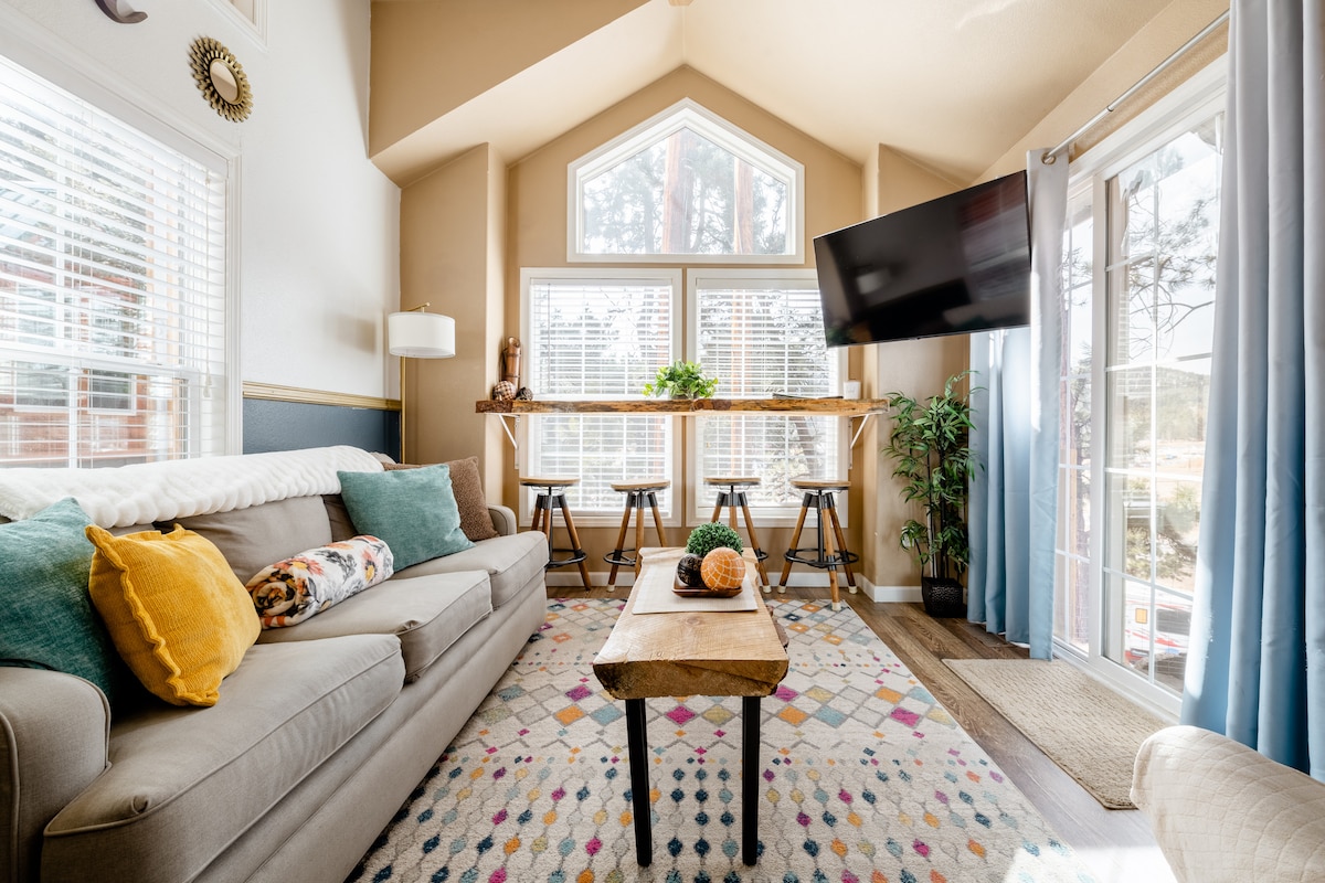 A bright living area features a comfortable gray sofa adorned with colorful cushions. A wooden coffee table sits in front, with a decorative item on top. Large windows allow natural light to fill the space, showcasing views of greenery outside. A flat-screen TV is mounted on the wall.