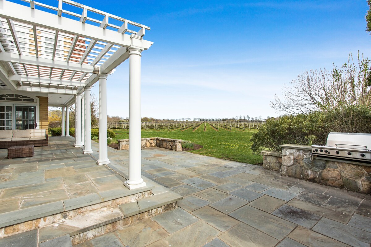 An outdoor patio area features stone flooring and is framed by white columns. A barbecue grill sits against a stone wall. Expansive views of lush vineyards extend into the distance, creating a serene backdrop under a clear blue sky.