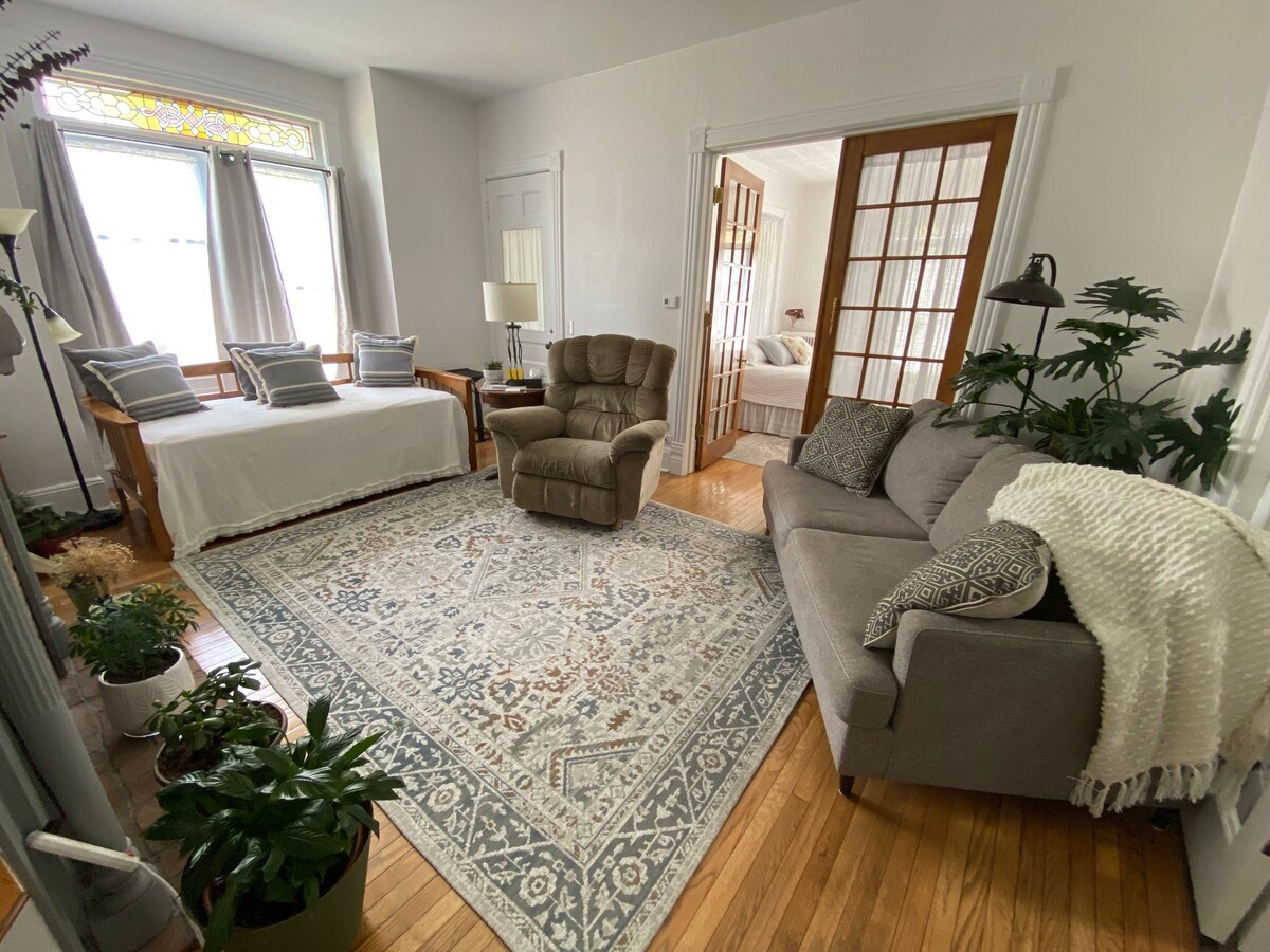 A spacious living area is highlighted by large windows that allow natural light to fill the room. A comfortable recliner and sofa are positioned around a decorative area rug, with various potted plants enhancing the inviting atmosphere. A daybed is visible towards the back, providing additional seating or sleeping space.