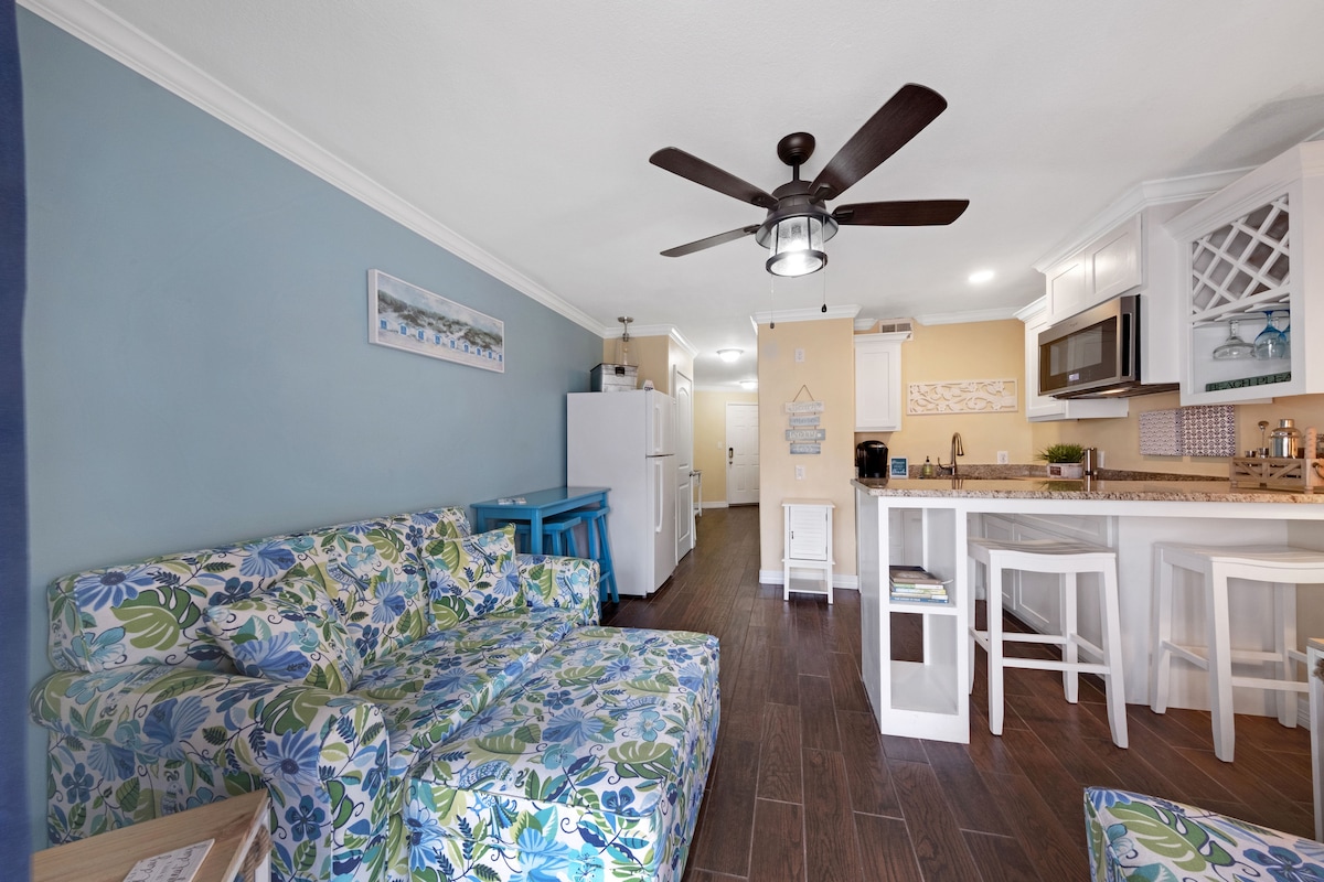 A modern living area is shown, featuring a floral-patterned sofa with a ceiling fan above. The space includes a kitchenette with a refrigerator, microwave, and a small dining table with four stools. Natural light brightens the room, highlighting the warm color scheme.