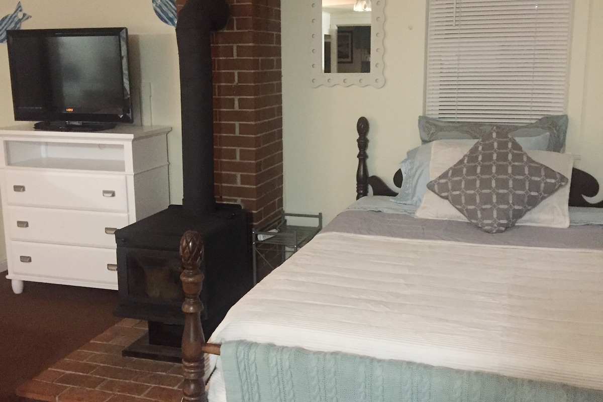 A cozy sleeping area features a spacious bed with light blue and white linens, complemented by a decorative pillow. A wooden dresser and a television are positioned nearby, while a wood stove with a brick surround adds warmth to the setting.