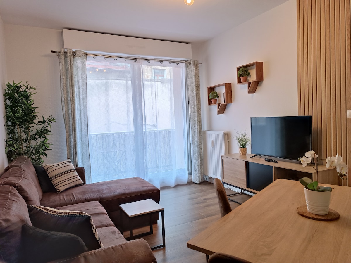 A cozy living area is presented, featuring a sectional sofa and a wooden coffee table. A mounted flat-screen television is visible on a light wood console. Natural light filters through sheer curtains, and decorative plants are arranged on wall-mounted shelves, adding a touch of greenery.
