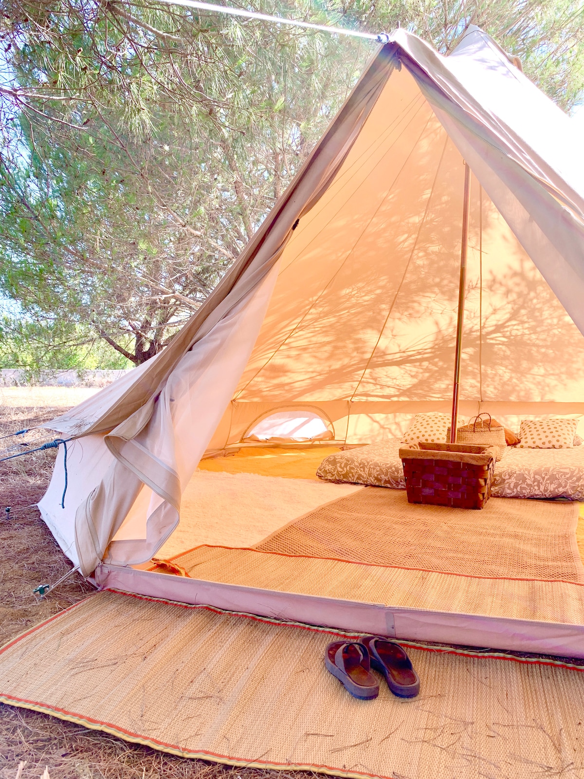 The interior of a spacious luxury tent is shown, featuring natural light filtering through the fabric and casting subtle shadows. Soft cushions are arranged on woven rugs, while a rustic basket sits on the floor. Simple footwear is placed outside the entrance.