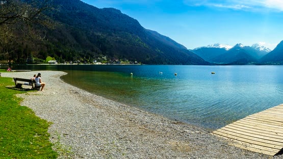 A serene lakeside view is captured, featuring clear water gently lapping at a pebbled shoreline. In the background, mountains rise against a blue sky, while a wooden dock extends into the lake. A person sits on a bench, enjoying the peaceful scenery.