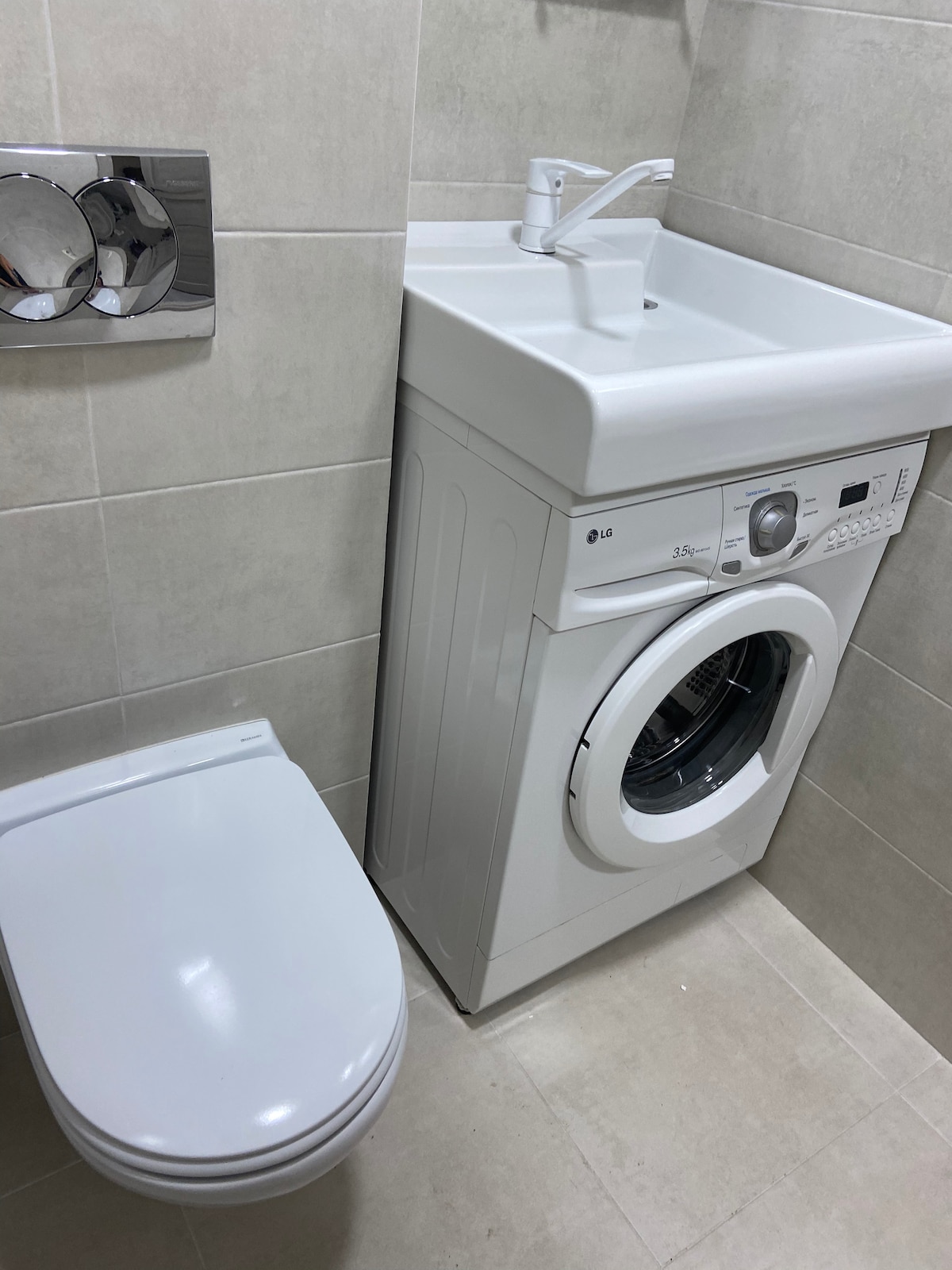 A compact bathroom space features a white toilet adjacent to a washing machine. The washing machine displays a control panel on its front, with a circular door for loading laundry. Light-colored tiles cover the walls and floor, creating a clean and modern look.