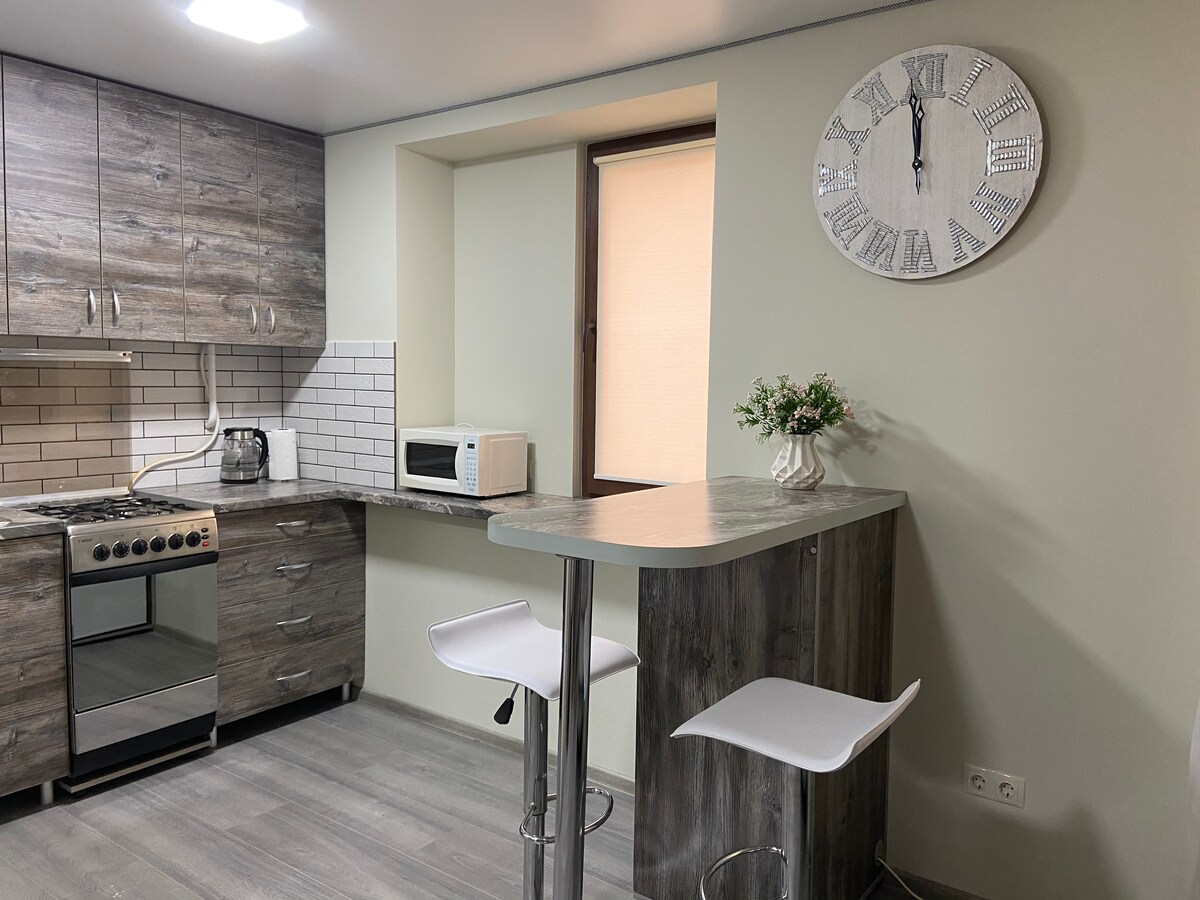 A modern kitchen area features a streamlined design with wooden cabinetry and white tiles. A small table with two bar stools is positioned next to a window. A microwave and kettle are visible on the countertop, while a large clock adorns the wall.