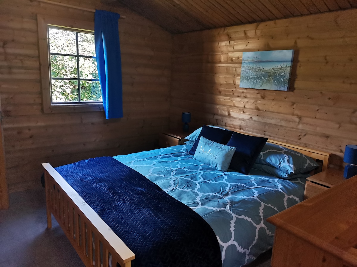 The sleeping area features a spacious bed adorned with a decorative blue and white quilt. Wooden walls create a rustic ambiance, complemented by a window with blue curtains allowing natural light. Nightstands with lamps are positioned on either side of the bed.