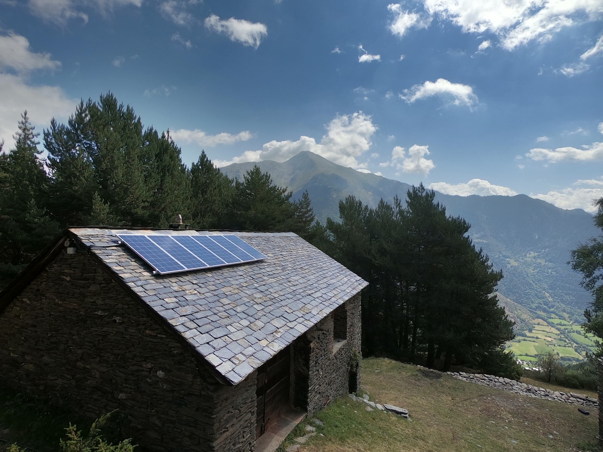A stone cabin is nestled among trees, featuring a slate roof with solar panels. The surrounding landscape includes rolling mountains and valleys under a partly cloudy sky, emphasizing the serene natural setting.