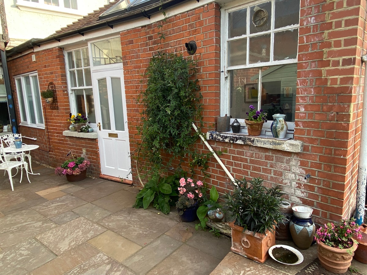 The external area features a charming brick wall with large windows. A white door leads to the interior. Potted plants with vibrant flowers are positioned along the pathway, contributing to a welcoming ambiance for outdoor enjoyment.