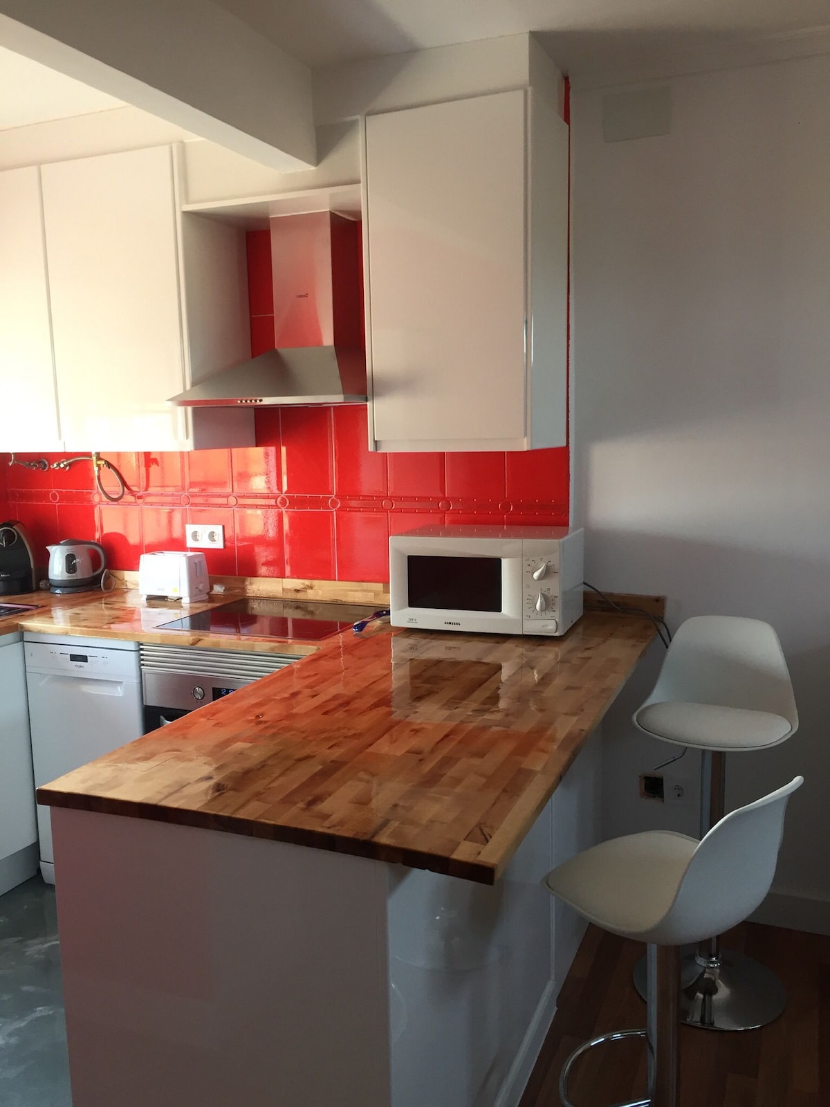 The kitchen features sleek white cabinetry and a vibrant red backsplash. A wooden counter provides seating for two on modern stools. Essential appliances, including a microwave and kettle, are arranged for convenience, complemented by a built-in dishwasher and stove.