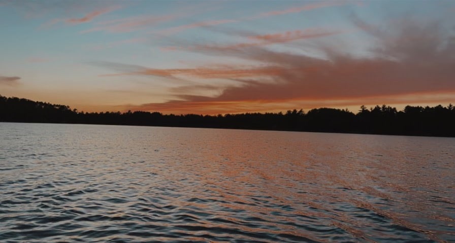 The serene waters of Sunset Lake reflect a vibrant sunset, with hues of orange and pink blending into a soft blue sky. The surrounding tree line creates a peaceful backdrop, emphasizing the tranquility of the evening atmosphere.