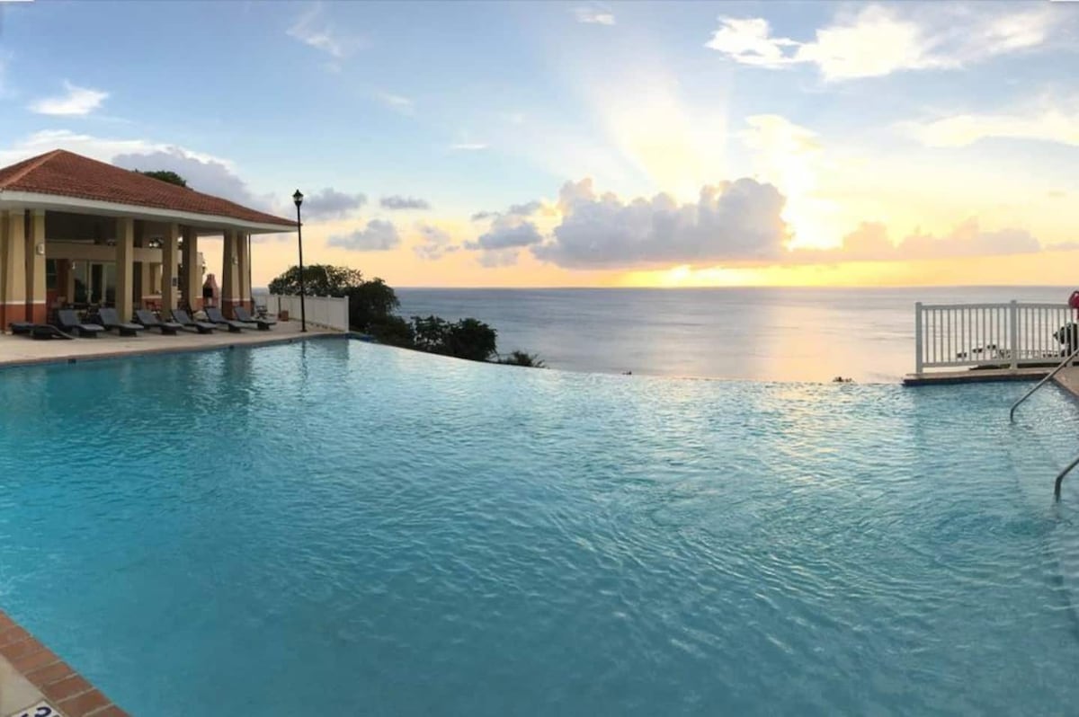 An expansive infinity pool reflects the vibrant colors of a sunset over the ocean. Cozy lounge chairs line the poolside, inviting relaxation. In the background, a covered outdoor patio provides shade and seating, enhancing the serene environment.