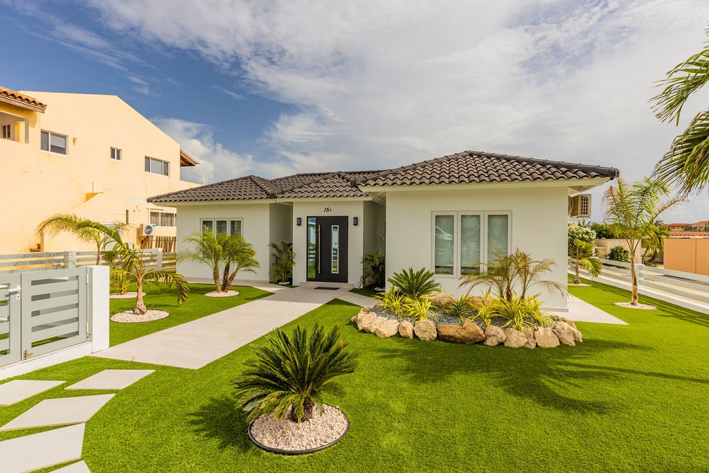 A modern villa is set within a lush green lawn, featuring a sleek pathway lined with decorative stones. Palm trees and native plants frame the entrance, while large windows are visible, enhancing the airy feel of the home's exterior.