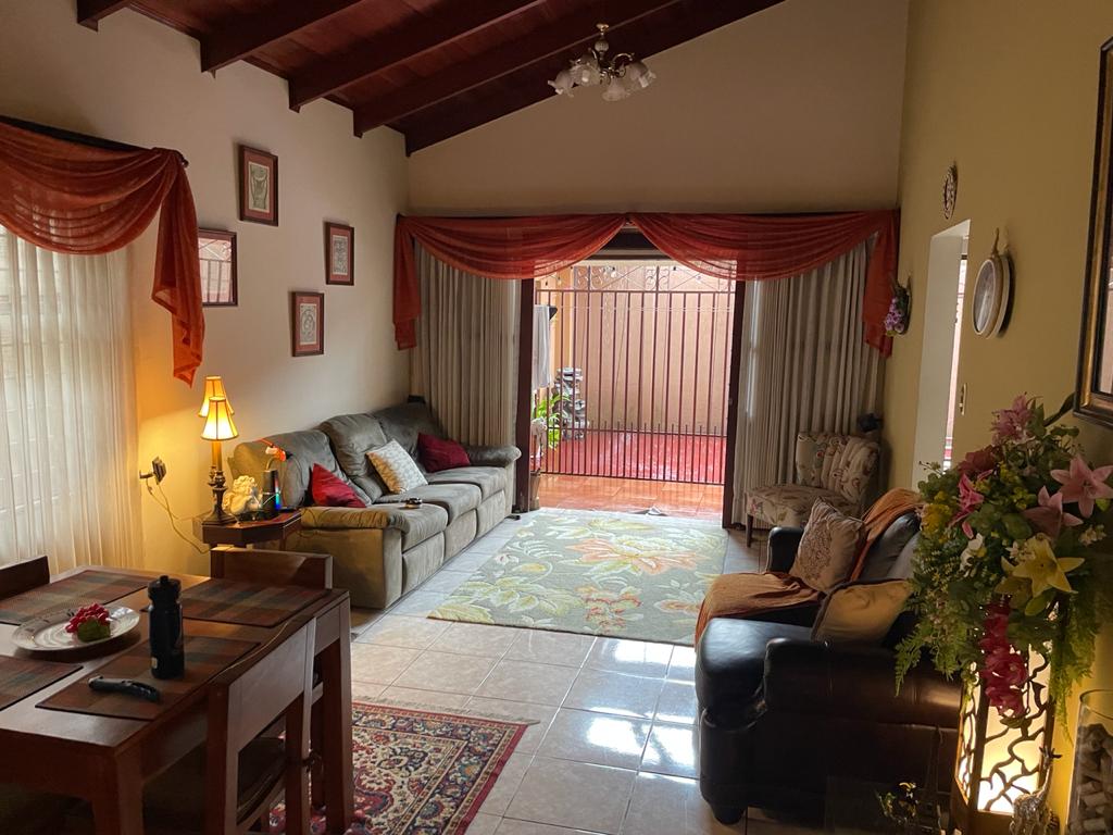 The living area showcases a mix of comfortable seating options, including a brown leather couch and a fabric sofa. Natural light filters through the sheer curtains, highlighting a patterned rug on the tiled floor. Decorative wall art and potted plants add warmth to the space.