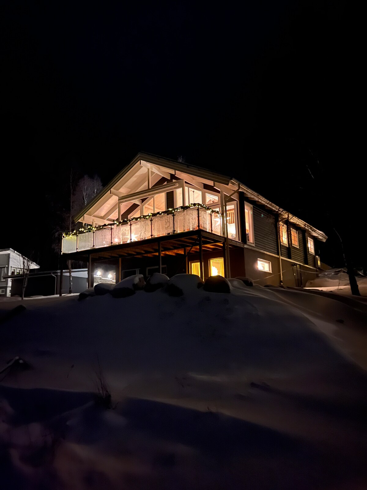 The villa is illuminated at night, showcasing a spacious balcony with inviting lights. The surrounding snow-covered landscape enhances the structure's charm, while large windows provide glimpses of the warm interior. The design features a high, sloping roof and tasteful outdoor decor.