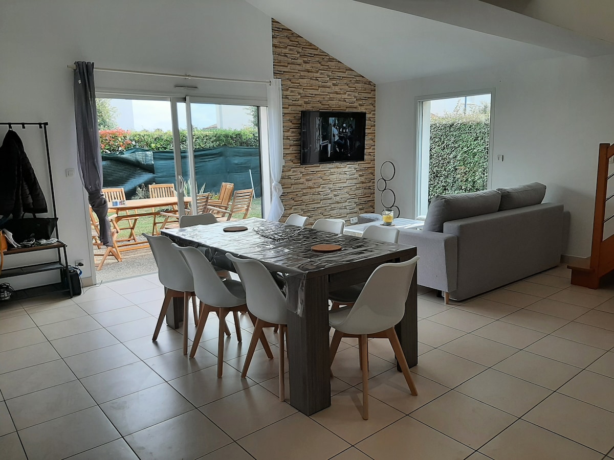 A spacious living area features large glass doors that open to an outdoor seating area. A wooden dining table with white chairs occupies the center of the room. A comfortable sofa is positioned against a textured stone wall, while a television is mounted for viewing.