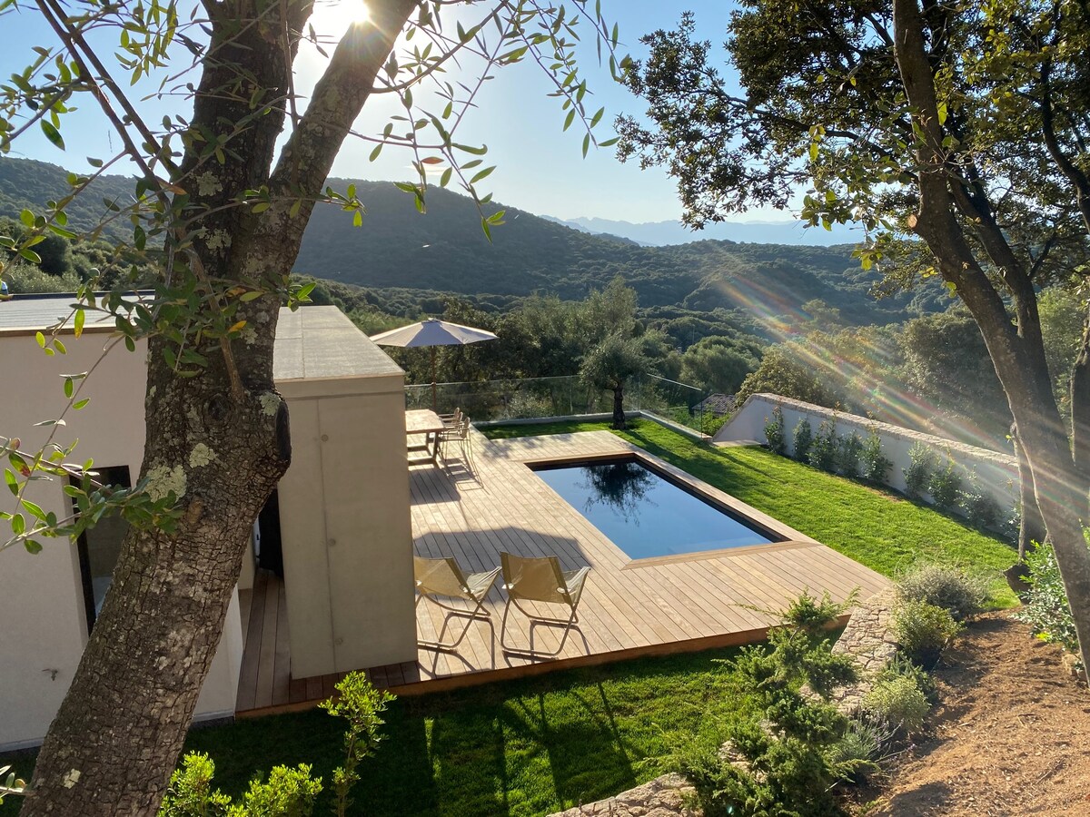 A modern villa is surrounded by lush greenery, showcasing a wooden deck with a private pool. Sunlight filters through the trees, creating a serene atmosphere. Two lounge chairs are positioned beside the pool, offering a peaceful outdoor space with mountain views in the background.