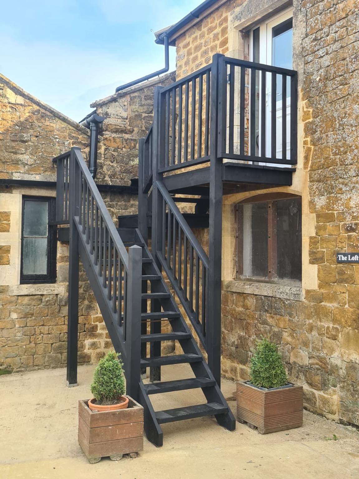 A set of dark wooden stairs ascends to the entrance of The Loft, flanked by two small potted plants. The surroundings feature rustic Cotswold stone walls and a glimpse of nearby windows, accentuating the traditional character of the building.