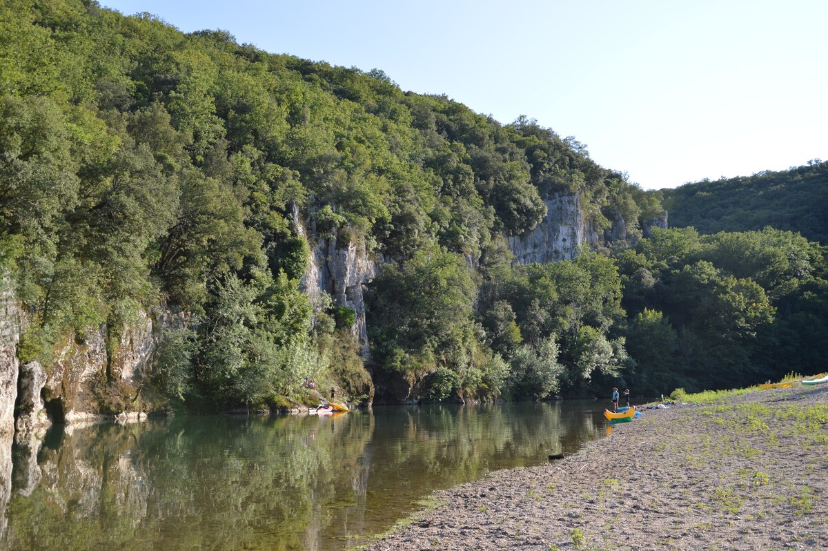 A serene river scene is displayed, with clear water reflecting surrounding greenery. The shoreline features a sandy area leading to lush trees and steep cliffs. Two kayakers can be seen enjoying the calm waters, emphasizing the natural beauty of the environment.