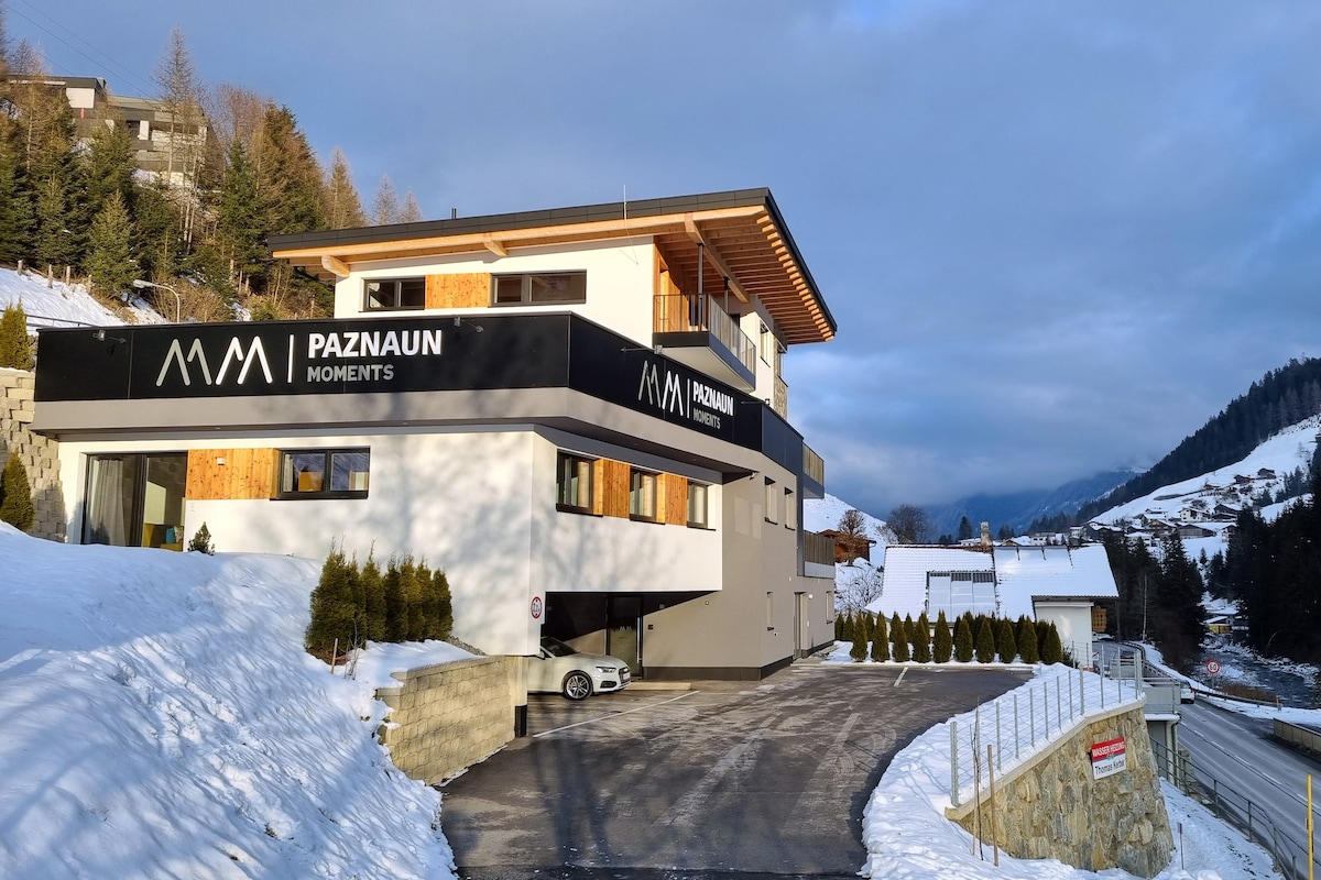 The modern building of Paznaun Moments is set against a snowy landscape. The structure features multiple levels with large windows and a covered entryway. Snow-covered shrubs line the pathway leading to the entrance, and the surrounding mountains are visible in the background.